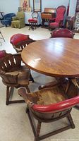 Round wooden dining table with six barrel back wooden chairs featuring red nailhead leather-type top upholstery in a room with other furniture items.