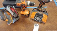 Photo showing Ridgid cordless circular saw, two cordless drivers, batteries, and battery charger on a brown surface.