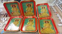 Six Coca-Cola trays showing vintage advertisement with woman in yellow dress and hat, arranged together.