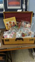 Open tan canvas suitcase showing interior red fabric pocket and numerous resealable bags of paper dolls and a Shirley Temple booklet