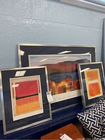 Three framed pieces of wall art arranged on a surface against a painted brick wall. One large Georgia O'Keeffe print centered with two smaller Mark Rothko prints on each side. Frames have silver metal edges and dark blue mats. Photos show visible scratches and smudging on the metal frames.