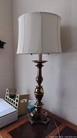 Full view of the brass table lamp on a wooden surface, showing the white fabric shade and the base with its detailed turned shape.