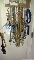 Multiple costume necklaces hanging on hooks against a wall with a visible lot tag.