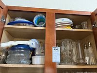 Two wooden cabinet shelves filled with assorted glassware including bowls, plates, candleholders, and salt and pepper shakers.