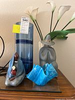 Photo showing tall blue glass vase with blue band, four white calla lilies, two ceramic figures, and blue abstract glass bowl on table against wall.