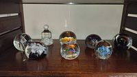 Photo of eight assorted glass paperweights arranged on wood surface, showing various internal colorful and abstract designs.