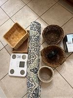 Top-down photo showing all items: baskets of different shapes and materials, rolled patterned bath rug, white digital scale with display, and metal scale.