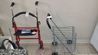Wheeled wire shopping cart and red Drive walker side by side on tile floor with wall background