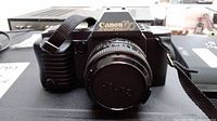 Front view of Canon T70 camera with lens cap attached on 50mm lens, showing camera body and strap.