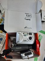 Lot of three cameras inside an open white box, including a Kodak EasyShare CX6200, Panasonic video camera with strap, and a red Canon camera.
