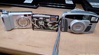 Three cameras displayed on a surface: from left to right Fujifilm FinePix E550 (silver), Olympus Stylus Tough-6000 (camouflage design), Fujifilm Discovery 290S (silver).