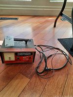 Front view of Mor Power Battery Charger Deluxe unit showing label and power switch, on a wood floor with attached cord.