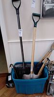 Plastic shovel, two wood-handled metal shovels, and two crowbars inside a blue bin showing wear and surface rust. Bin not included.