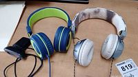 Photo showing blue and green MPow headphones and white Marley headphones with snake skin headband on a wooden surface, with visible cables and a clear Logitech webcam attached to a mount.