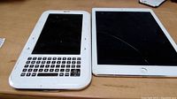 Photo showing white Ebook reader with physical keyboard next to white iPad with cracked screen.