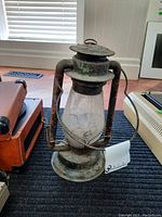 Front view of heavily worn metal lantern with glass globe placed on a dark rug with vintage items nearby.
