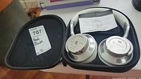 Black zippered carrying case open, holding silver Paww wireless headphones with warranty paper inside