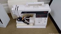 White Elna 6000 sewing machine set on carpet with some sewing accessories resting on the base, front view.