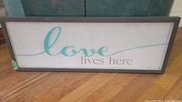 Front view of the rectangular wood framed wall art with the phrase "love lives here" in teal and gray on white.