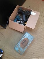 Cardboard box with assorted hand tools and power tools including a black and blue Bosch drill among other industrial drills with cords inside.