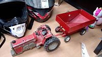 Photo showing MF 2775 tractor toy alongside a red trailer and two helmets on a flat surface, highlighting the missing rear wheel on the tractor.