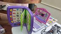 Purple Barbie B-Book electronic learning tool with alphabet pages open and visible, placed next to pink Barbie toy laptop and puzzles on table.
