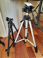 Two adjustable camera tripods side by side on indoor wooden floor, one silver-black and one all black, showing size difference and mounting heads with handles.