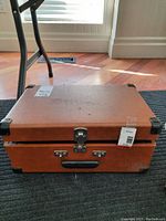 Brown suitcase-style carrying case closed, showing metal clasps, black handle, minor surface wear.