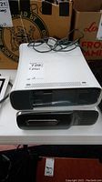 Two white Xbox 360 consoles stacked on a table with a partial power cord visible on top console. Signs of scratches and wear present.