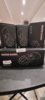 Four boxed sets of battery powered rechargeable heated gloves placed on a surface, boxes show graphic outlines of gloves and heating indicators.