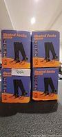 Four orange and blue boxed retail packages of battery powered rechargeable heated socks stacked in a 2x2 layout on a black surface under ceiling lights.