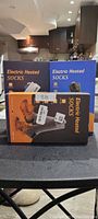 Photo showing three boxes of electric heated socks stacked on a table, two blue and one orange box, unopened and labeled.