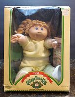 Doll in original windowed yellow and green box labeled 1984 Cabbage Patch Kids with doll visible inside dressed in pale yellow outfit