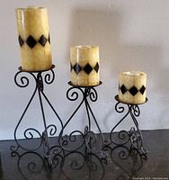 Three wrought iron candle holders with intricate scrollwork and pillar-style tealight candles placed on them, varying in height and size.