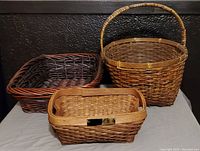 Front angled photo showing all three darker wicker baskets of varying sizes and styles, one large rectangular, one oval with handle, and one smaller square with handle and metal plate