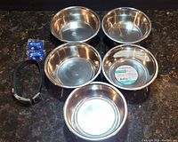 Five Maslow stainless steel dog bowls arranged together showing their size and non-skid bases with small dog collar and two blue flashing safety lights.