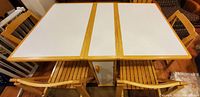 View of dining table with drop leafs open, showing white laminate surface bordered with natural wood frame and two folding chairs on each side.