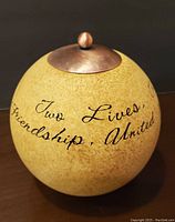 Side view showing the text 'Two Lives, Friendship, United' on the beige spherical tealight candle holder with copper lid.