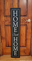 Front view of the vertical 'Home Sweet Home' sign leaning against a wooden door showing the design and colors.