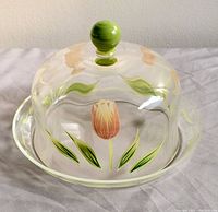 Clear crystal two-piece butter dish with dome lid and base plate, featuring hand-painted pink tulip and green leaves, dome handle painted green, displayed on white cloth background.