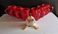 Front view showing six red mugs arranged in two rows with the small rooster figurine in front.