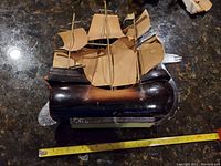 Photo of a wooden ship model with three masts, brown wooden hull, and fabric sails. The bow features a hand-shaped decoration. Sails and rigging appear aged and some sails torn or creased. Measuring tape shows approximately 20 inches in length.