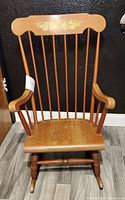 Front view of vintage rocking chair showing vertical spindles, floral motif on top backrest, curved arms, and cracked seat.