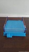 Blue plastic wrestling ring playset on a dark wood surface, showing front and bottom sides with four blue ring posts and three ropes in red, white, and blue.