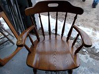 Close-up image of an oak captain's chair showing the contoured wooden seat, spindle back with cutout handle, and curved armrests.