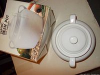 Top view of white ceramic bean pot with lid beside the original box showing product photo and description.