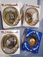 Four M.J. Hummel branded Christmas tree ornaments in original boxes, three white and one blue "Charming Classics" box, showing front painted scenes with gold bead trim on glass ornaments.