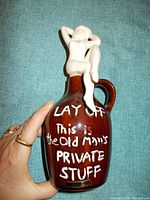 Brown ceramic jug labeled 'Old Man's Private Stuff' with a small figurine seated on top, held in hand.