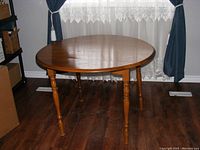 Photo of the round oak kitchen table with drop-leaves fully up, showing the natural wood grain and warm finish, standing on dark wood flooring with a window and blue curtains in the background.
