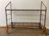 Front view of the two-tier brown metal wire shelf with wooden legs against a plain wall and carpet floor.